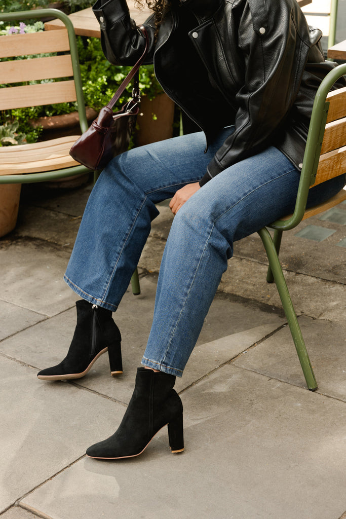 Person sitting on a chair wearing black leather jacket, blue jeans, and black ankle boots.