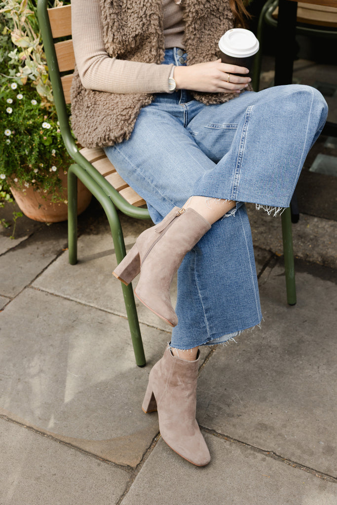 Person sitting outdoors holding a coffee cup, wearing jeans, taupe ankle boots and a vest.
