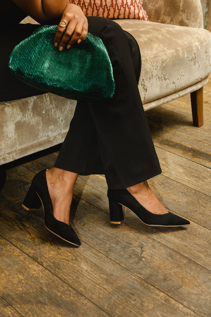 Person sitting on a couch holding a green clutch, wearing black high-heeled shoes.