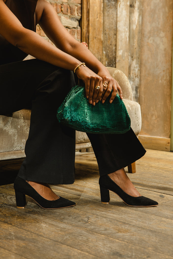 Person wearing black high-heeled shoes and holding a green clutch in a rustic setting.