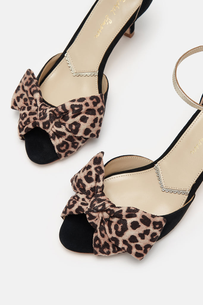 close up detail shot of a pair of black suede low heel two part occasion shoes, with oversized leopard bows and gold leather ankle straps