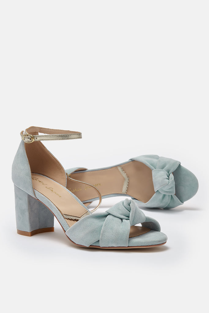pair of dusky blue suede block heel shoes, with knot detail and gold ankle strap- one shoe is lying on its side