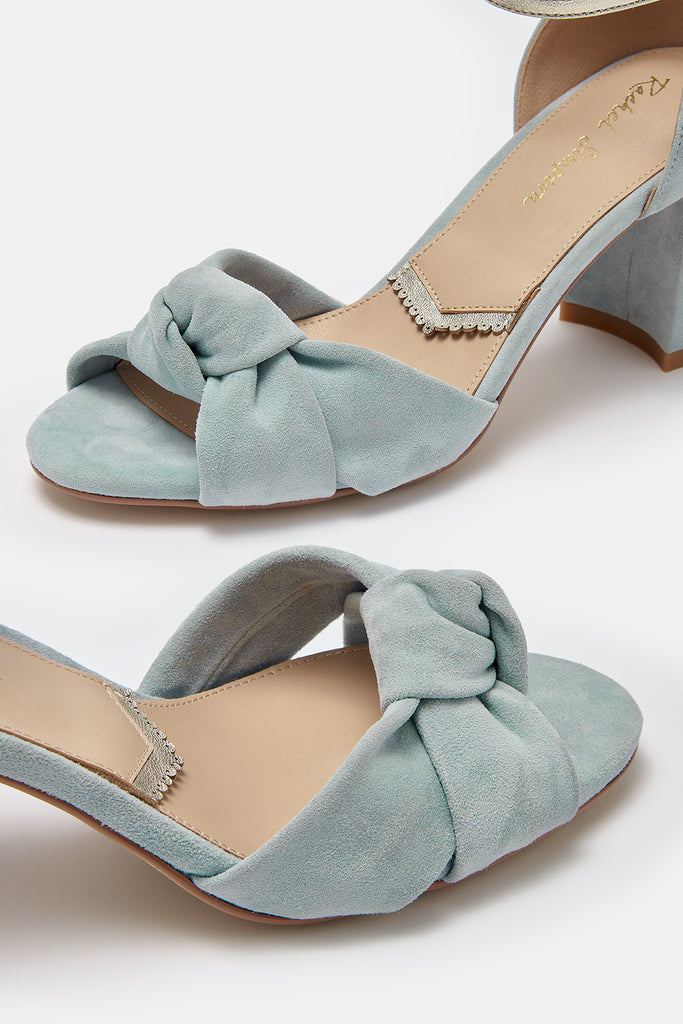 close up detail shot of a pair of dusky blue suede block heel shoes, with knot detail and gold ankle strap