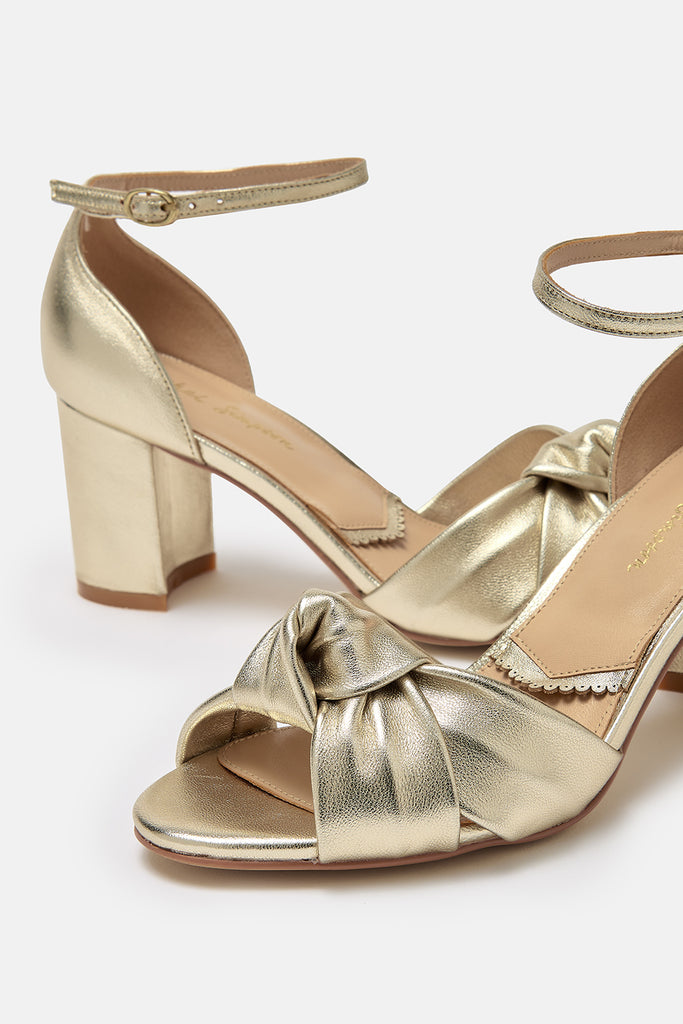 close up detail shot of a pair of gold leather block heel occasion shoes with knot detail and ankle straps