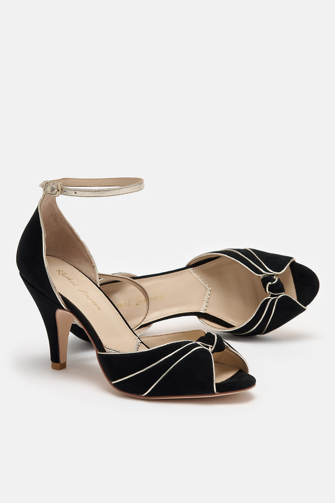 pair of black and gold suede occasion shoes with vintage inspired knot detail, mid heels and gold leather ankle straps- one shoe is lying on its side