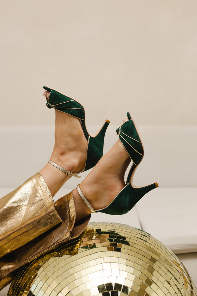 woman's legs in a pair of gold trousers, wearing a pair of forest green and gold two part occasion shoes, with vintage inspired knot detail, mid heels and gold leather ankle straps. Her feet are resting on a large gold disco ball