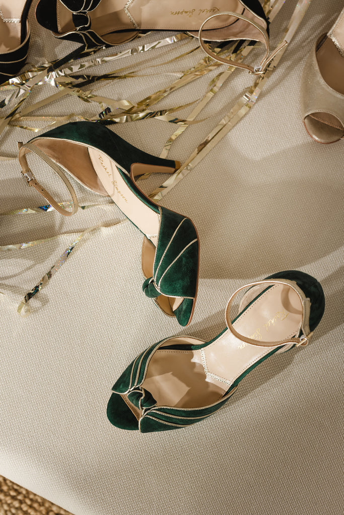 pair of forest green and gold two part occasion shoes, with vintage inspired knot detail, mid heels and gold leather ankle straps, on a cream surface surrounded by gold streamers
