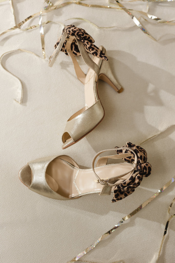 pair of antique gold leather ankle strap sandals with oversized leopard bows on the back and mid height heels, on a cream background surrounded by gold streamers