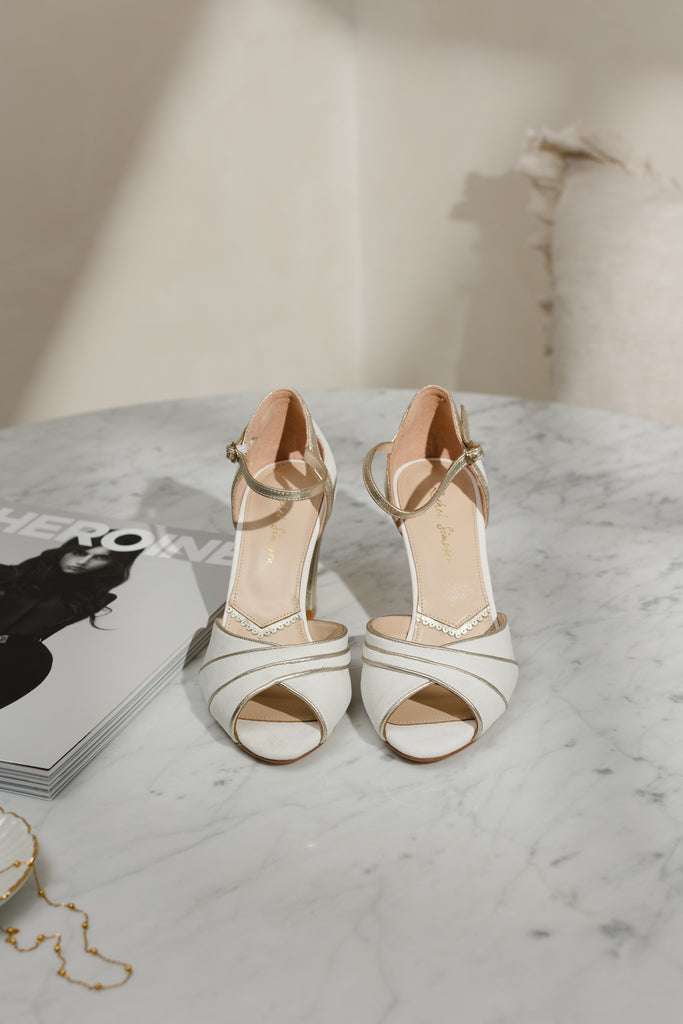 pair of ivory suede wedding shoes with gold block heel and ankle strap, on a marble surface next to a magazine and gold necklace