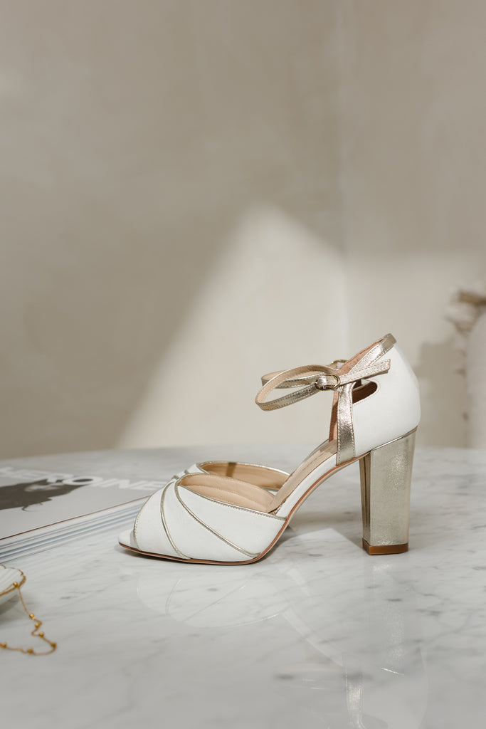 pair of ivory suede wedding shoes with gold block heel and ankle strap, on a marble surface next to a magazine and gold necklace- side view