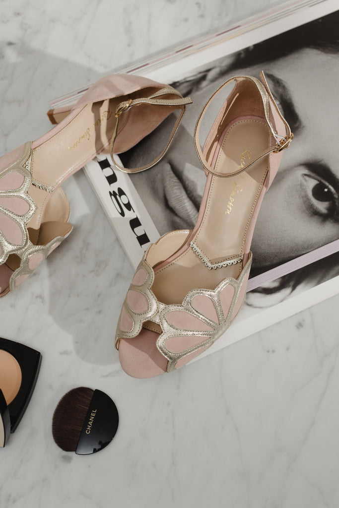 pair of blush suede suede and gold art deco vintage style shoes, with low heels and ankle strap, sitting on a magazine on a marble surface, next to a powder compact