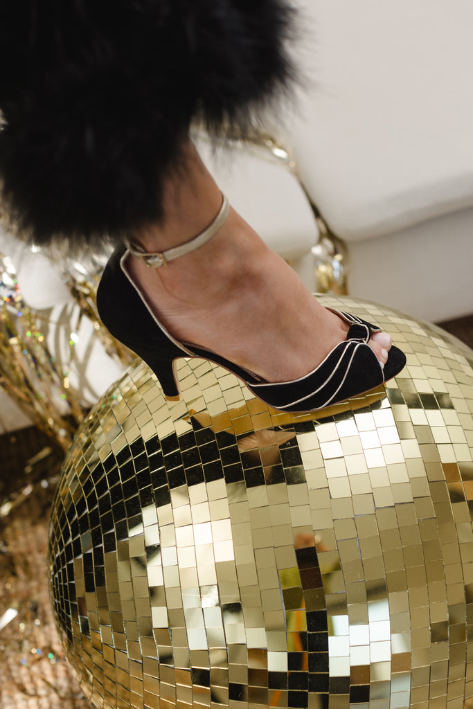 woman's foot resting on a gold disco ball, wearing a black and gold suede occasion shoe with vintage inspired knot detail, mid heel and gold leather ankle strap