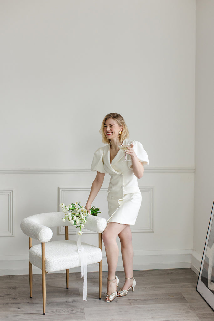 bride holding a glass of champagne and a bouquet of cream flowers, wearing a pair of ivory suede and gold art deco vintage style wedding shoes, with low heels and ankle strap