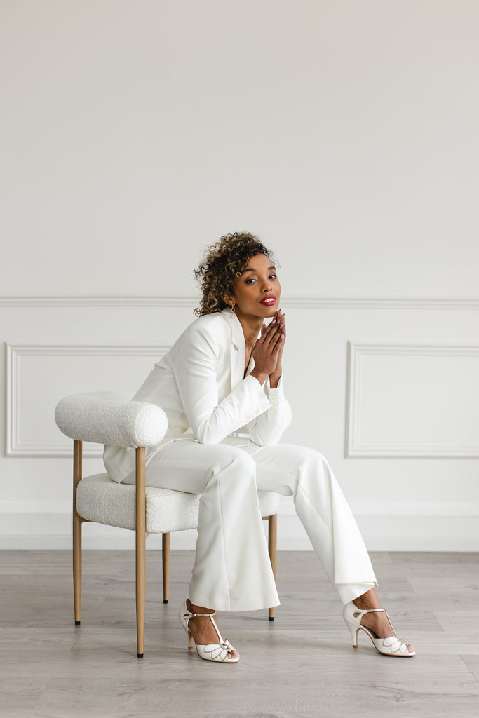 bride in an ivory trouser suit, sitting in a chair wearing a pair of pair of ivory leather and gold t-bar wedding shoes with mid heels and ankle strap