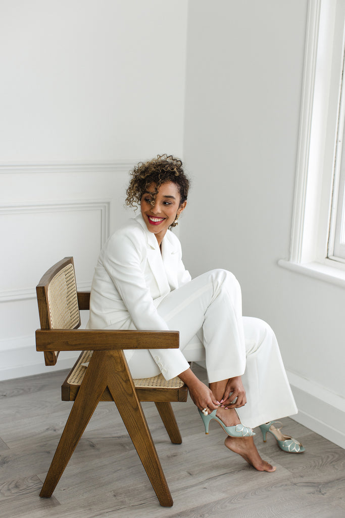 smiling model in an ivory trouser suit, sitting in a chair putting on a pair of mint green and gold leather t-bar occasion shoes with mid heels and ankle strap
