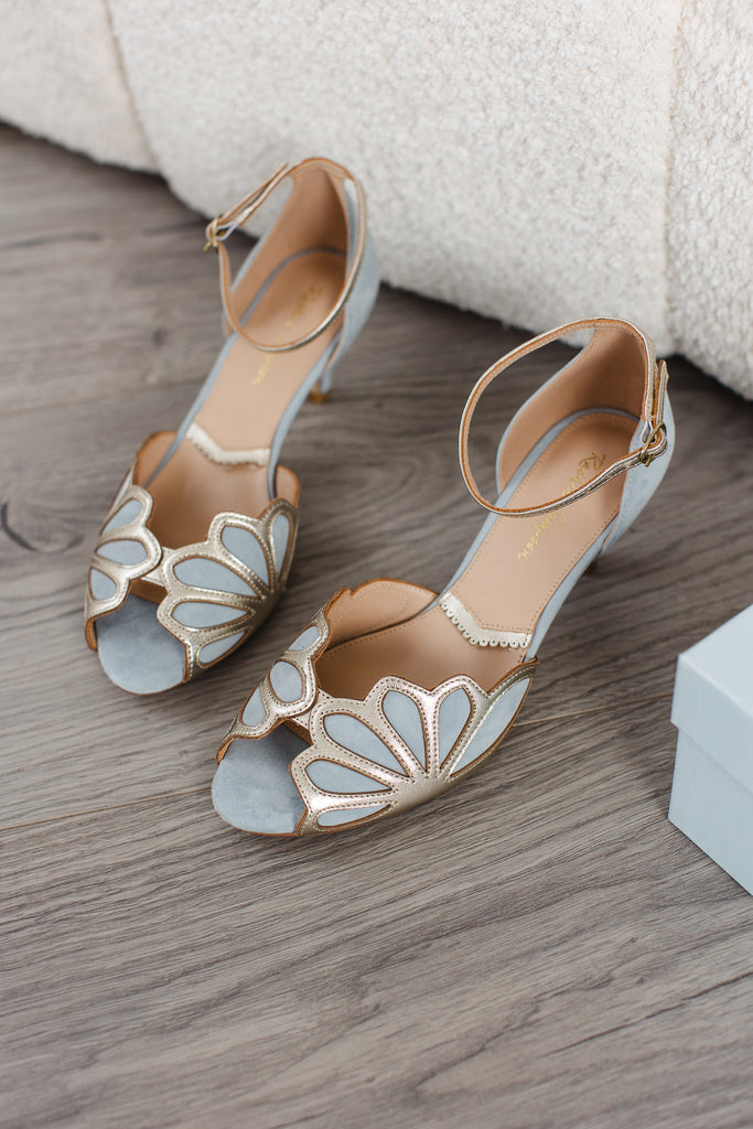 pair of dusky blue suede and gold art deco vintage style shoes, with low heels and ankle strap placed on a wooden floor, next to a shoe box