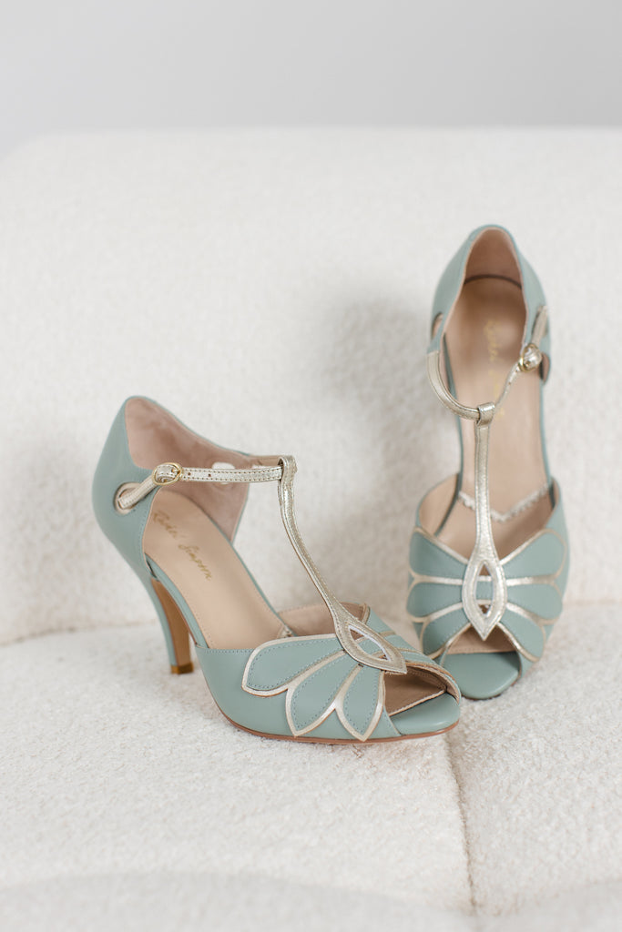 a pair of mint green and gold leather t-bar occasion shoes with mid heels and ankle strap, on a cream sofa