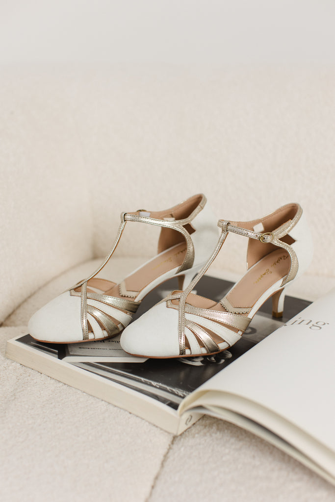 pair of ivory suede and gold leather twenties vintage style t-bar wedding shoes, with low heels and ankle straps, sitting on a magazine on a cream sofa
