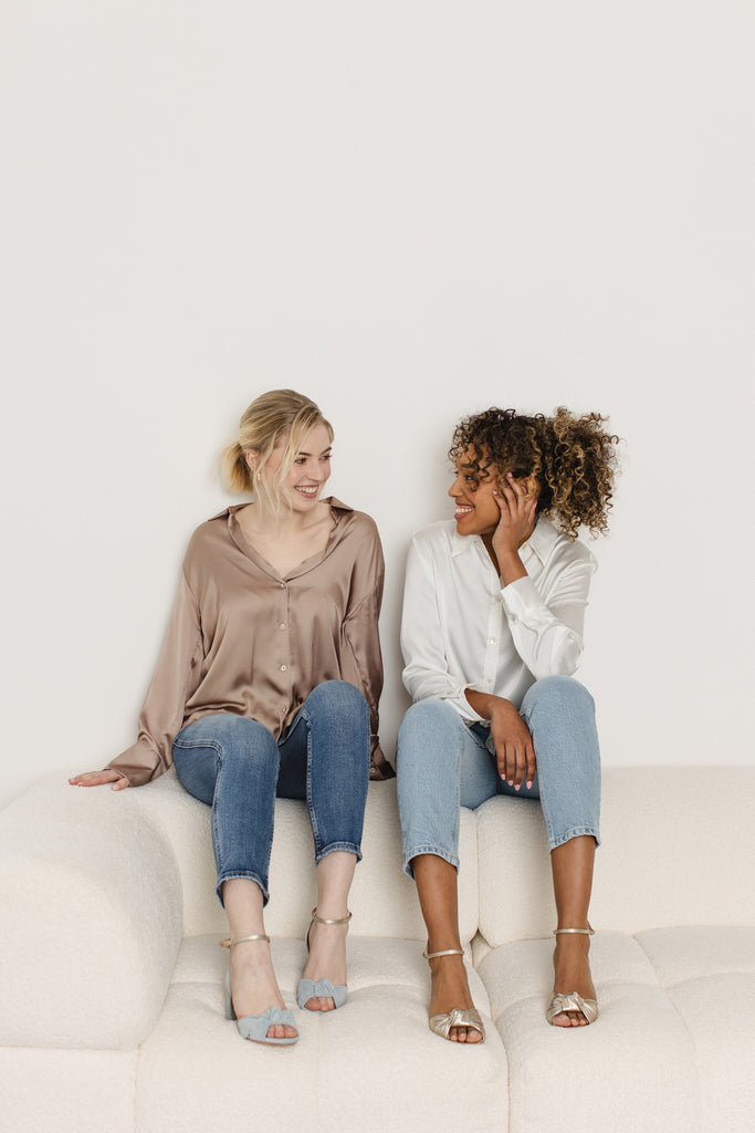 two smiling models sitting on a cream sofa- one is wearing a pair of dusky blue suede block heel shoes, with knot detail and gold ankle strap, the other is wearing a pair of gold leather block heel shoes with knot detail and ankle strap