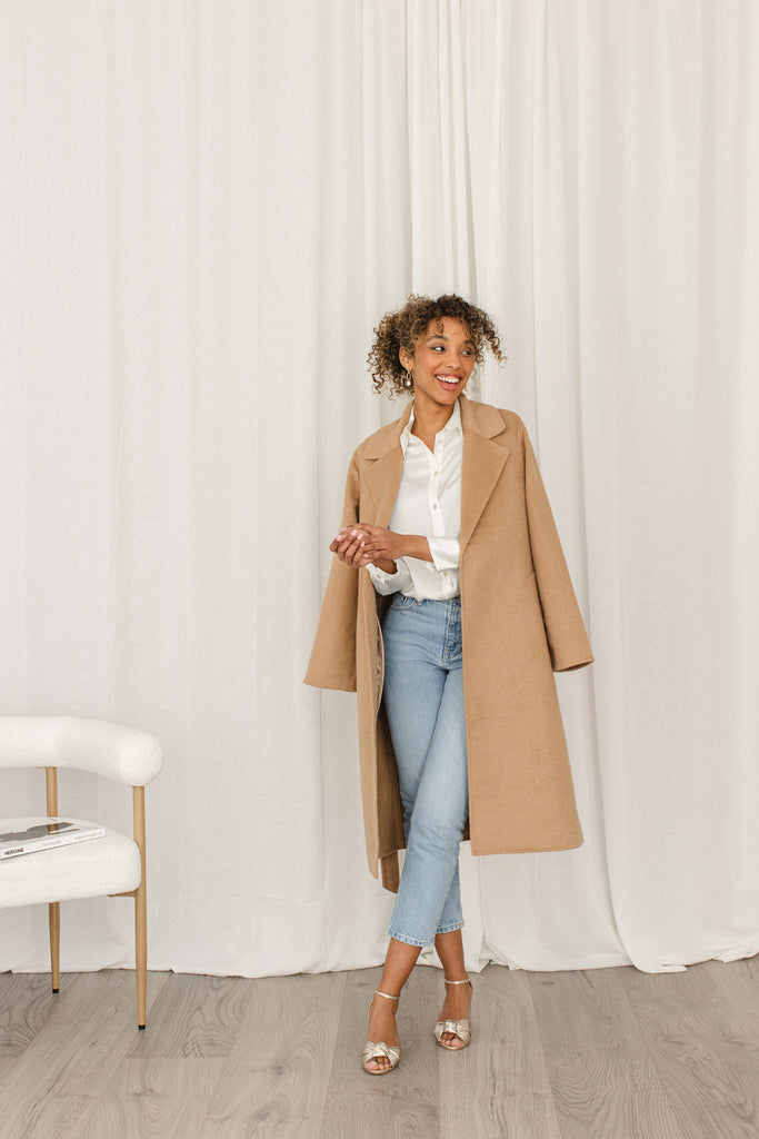 smiling woman in jeans and a camel coat, wearing a pair of gold leather block heel occasion shoes with knot detail and ankle straps