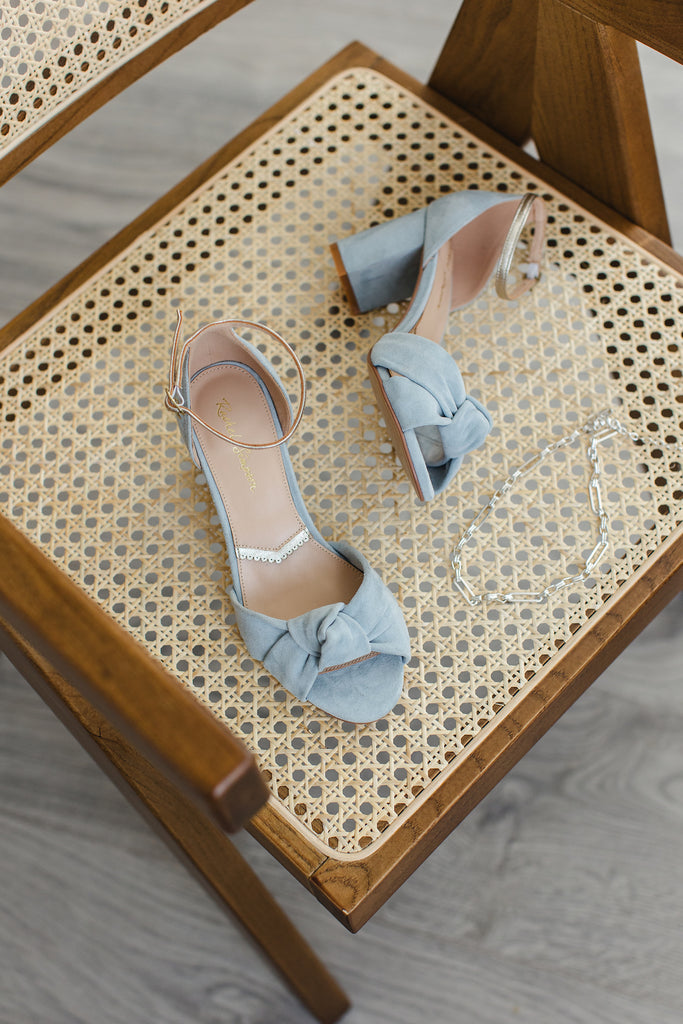 pair of dusky blue suede block heel shoes, with knot detail and gold ankle strap, placed on a chair next to a silver necklace