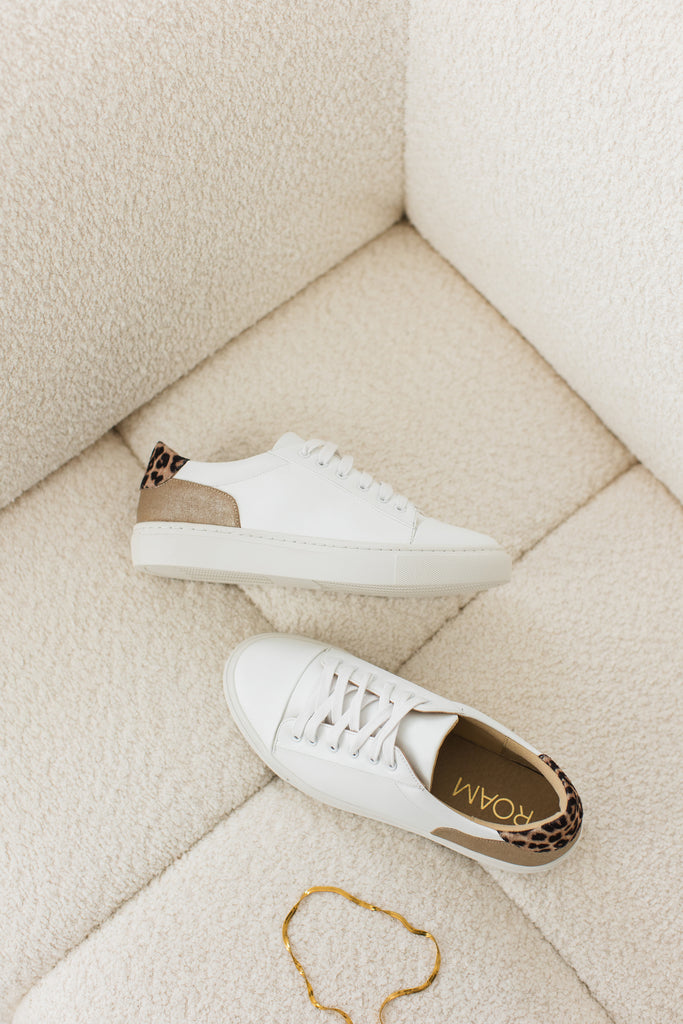 Pair of white vegan leather trainers with gold and leopard heel detail on a cream boucle sofa, with a gold necklace nearby