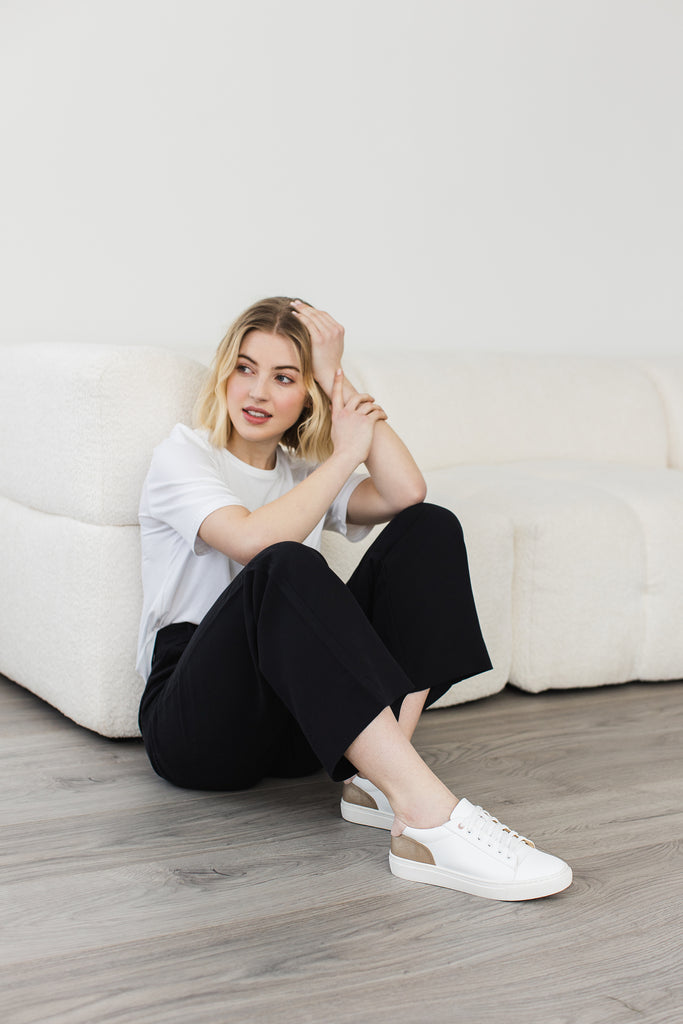 model sitting on floor wearing a pair of white vegan leather trainers with gold and pink heel detail