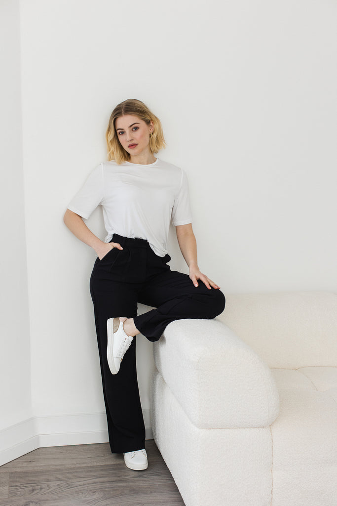 model standing against a wall wearing a pair of white vegan leather trainers with gold and pink heel detail, resting one leg on the arm of a cream boucle sofa