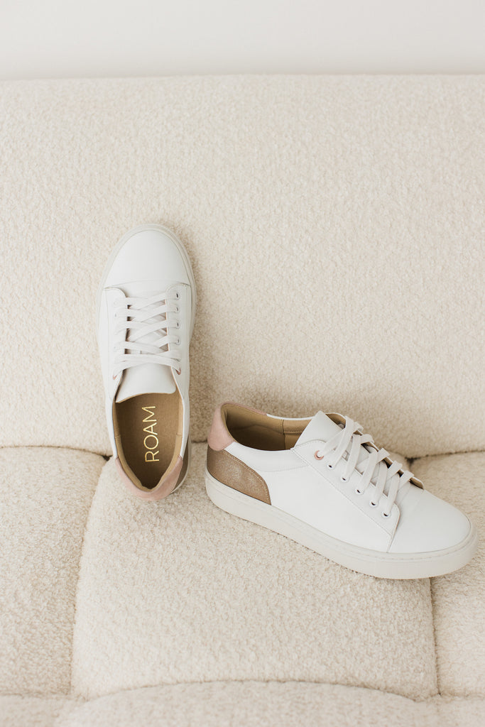 pair of white vegan leather trainers with gold and pink heel detail, placed on a cream boucle sofa