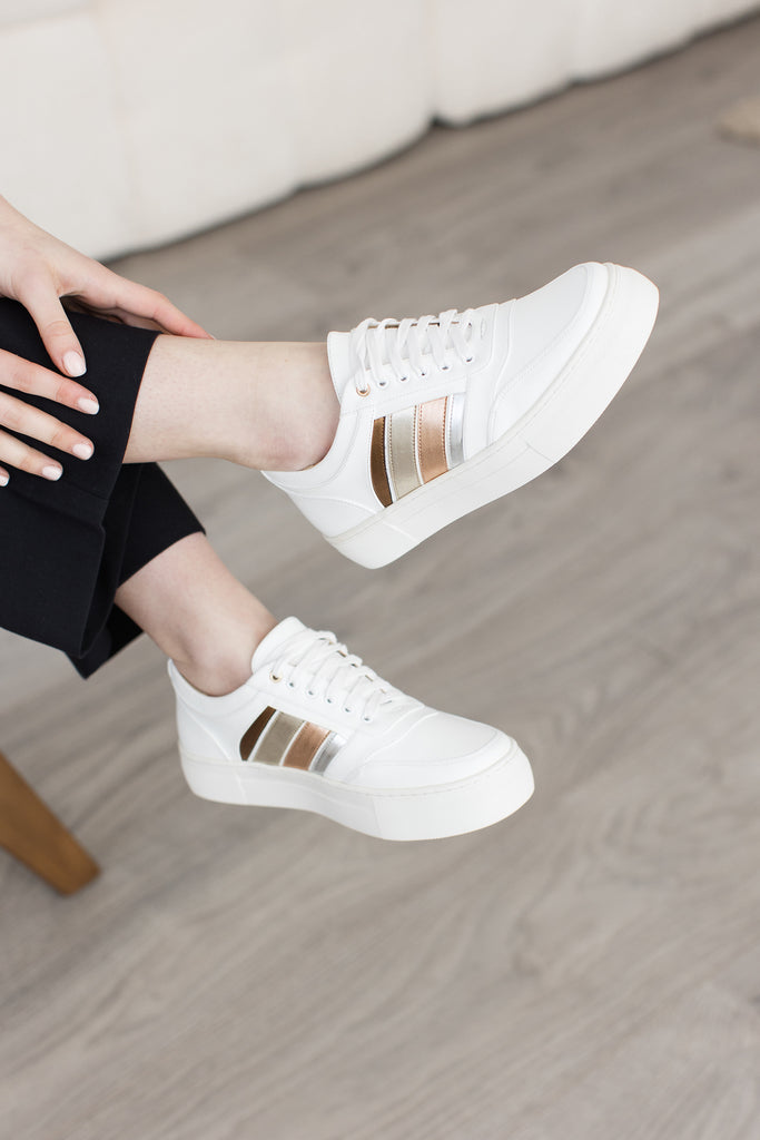 Close up foot shot of model wearing white vegan leather trainers with side stripes in bronze, gold, rose gold and silver.