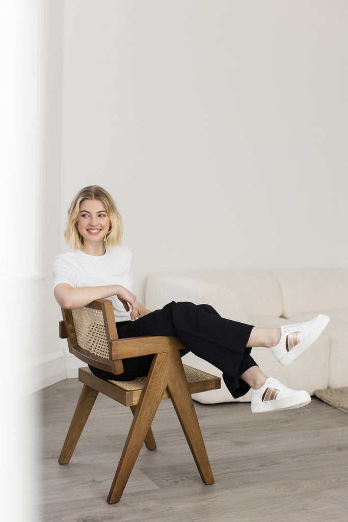 Smiling model sitting in a chair with her legs over one arm, wearing a pair of white vegan leather trainers with side stripes in bronze, gold, rose gold and silver.