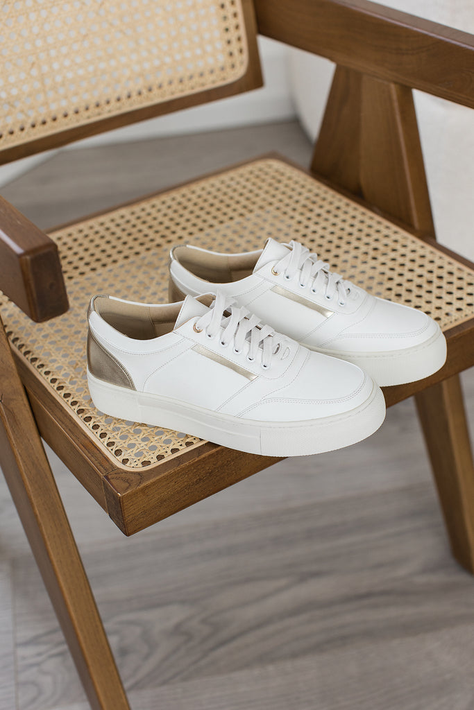 Pair of white and gold vegan leather trainers placed on a natural coloured chair.