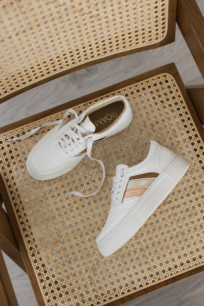 A pair of white vegan leather trainers with side stripes in bronze, gold, rose gold and silver, placed on a natural coloured chair.
