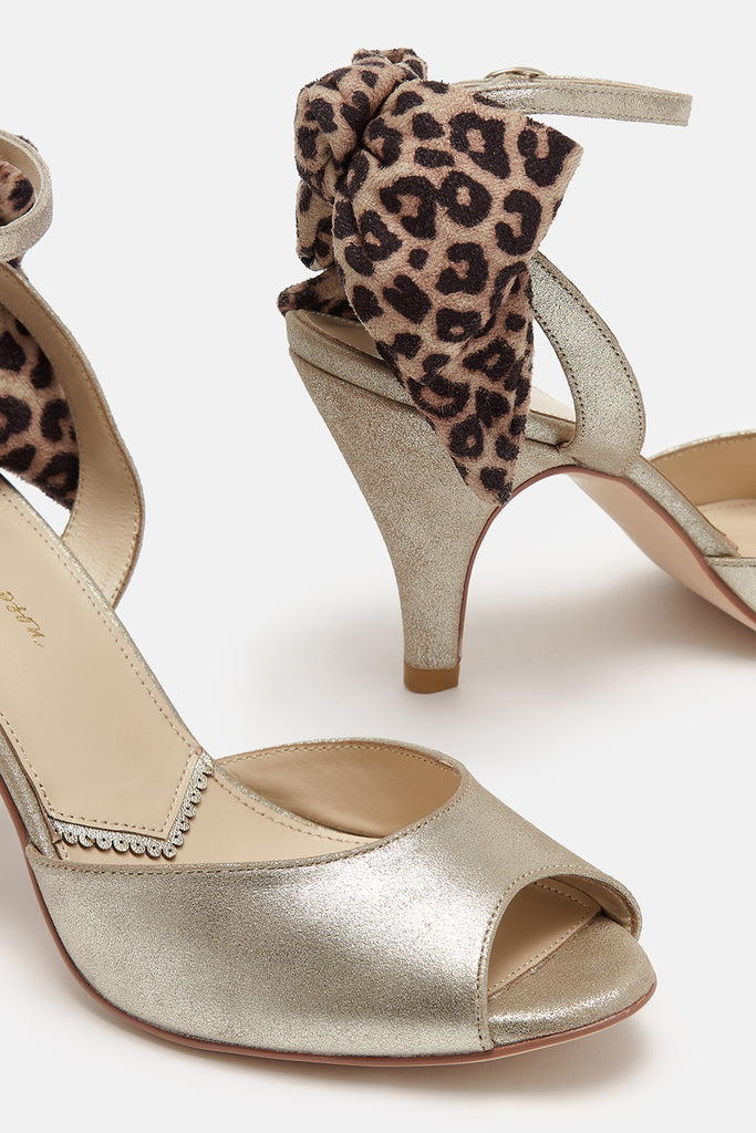 close up detail shot of a pair of antique gold leather ankle strap sandals with oversized leopard bows on the back and mid height heels